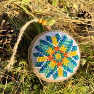 May include: A round, white, fabric ornament with a colorful geometric design in shades of blue, green, yellow, and orange. The ornament is hanging from a brown string.