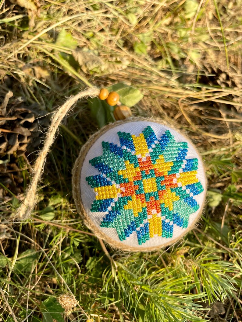 May include: A round, white, fabric ornament with a colorful geometric design in shades of blue, green, yellow, and orange. The ornament is hanging from a brown string.