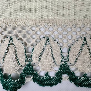 May include: Close-up of a cream-colored linen fabric with a decorative border. The border features a delicate openwork design with alternating cream and dark green crochet elements, creating a textured and detailed edge.