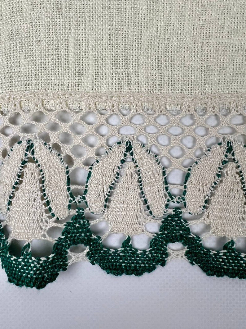 May include: Close-up of a cream-colored linen fabric with a decorative border. The border features a delicate openwork design with alternating cream and dark green crochet elements, creating a textured and detailed edge.