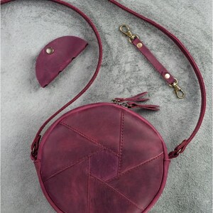 May include: A burgundy leather round crossbody bag with a matching coin purse and strap. The bag features a geometric design and a zipper closure with tassel pulls. The strap has gold-colored clasps.