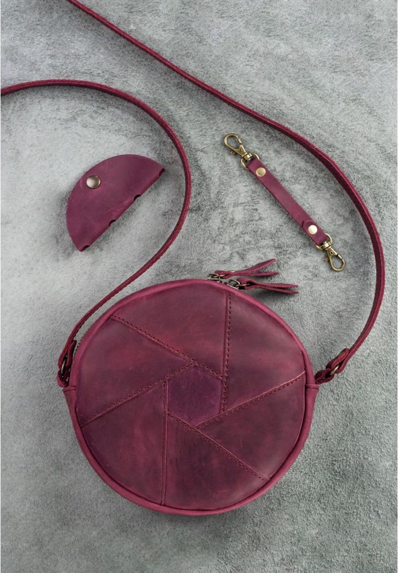 May include: A burgundy leather round crossbody bag with a matching coin purse and strap. The bag features a geometric design and a zipper closure with tassel pulls. The strap has gold-colored clasps.