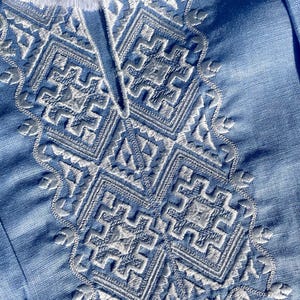 May include: Close-up of a light blue garment with intricate white embroidery. The embroidery features geometric patterns and floral accents around the neckline. White tassels are visible at the top of the garment.