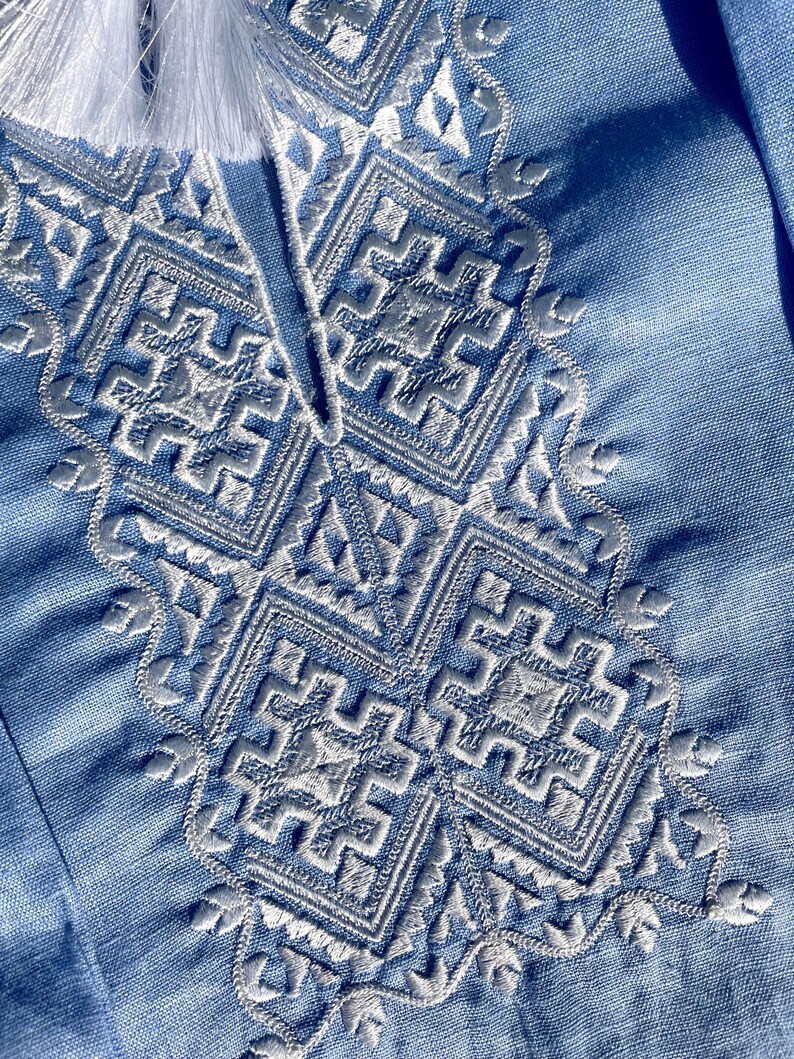 May include: Close-up of a light blue garment with intricate white embroidery. The embroidery features geometric patterns and floral accents around the neckline. White tassels are visible at the top of the garment.