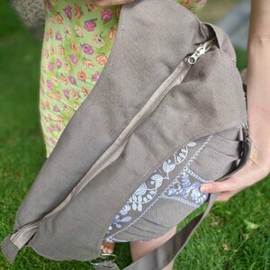Peut inclure: Un sac cabas en toile gris avec une fermeture &eacute;clair et un motif floral brod&eacute; blanc. Le sac a une longue bandouli&egrave;re et est port&eacute; &agrave; l'&eacute;paule.