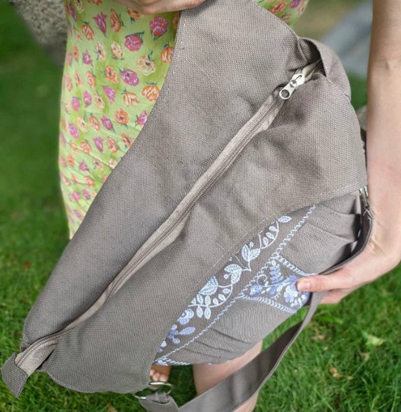 Peut inclure: Un sac cabas en toile gris avec une fermeture &eacute;clair et un motif floral brod&eacute; blanc. Le sac a une longue bandouli&egrave;re et est port&eacute; &agrave; l'&eacute;paule.