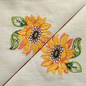 May include: Two embroidered sunflowers with green leaves on a white linen fabric. The sunflowers are yellow with orange centers and brown centers.
