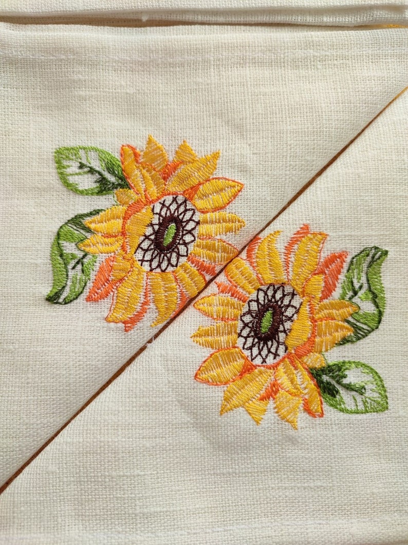 May include: Two embroidered sunflowers with green leaves on a white linen fabric. The sunflowers are yellow with orange centers and brown centers.