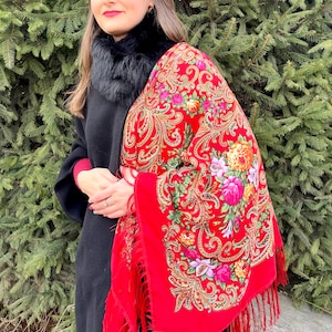 May include: A woman wearing a black coat and a red shawl with a floral pattern and fringe. The shawl is draped over her shoulders and has a black fur collar.