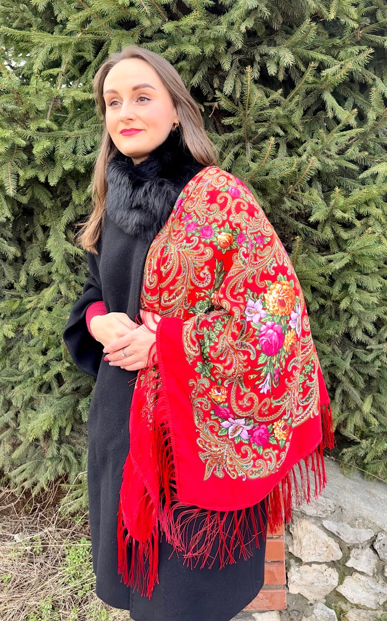 May include: A woman wearing a black coat and a red shawl with a floral pattern and fringe. The shawl is draped over her shoulders and has a black fur collar.
