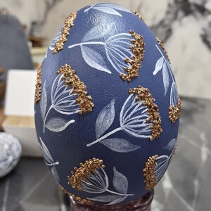 Puede incluir: Un huevo decorativo azul marino con dise&ntilde;os florales blancos y detalles dorados. El huevo presenta flores y hojas blancas pintadas a mano, con adornos dorados texturizados. Un art&iacute;culo decorativo &uacute;nico.