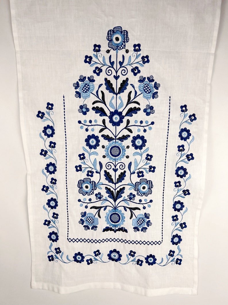May include: White linen fabric with blue embroidered floral design. The design features a central floral motif with smaller floral motifs along the sides.