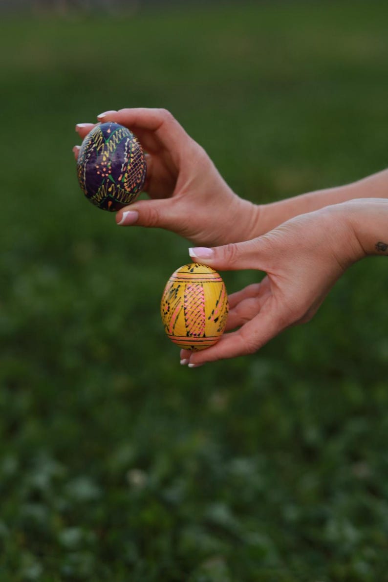 K&ouml;nnte beinhalten: Zwei bemalte Ostereier, eines lila mit goldenen und gr&uuml;nen Mustern und eines gelbes mit rosa und schwarzen Mustern, werden in den H&auml;nden einer Person gehalten.