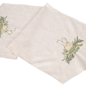 May include: Two beige linen table runners with an embroidered design of a green olive branch and a clear glass bottle.