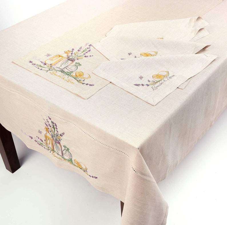 May include: A beige linen tablecloth and matching napkins are displayed on a wooden table. The tablecloth and napkins feature embroidered designs of lavender, lemons, and jars. The text "Lavender & Lemon" is also embroidered on the tablecloth and napkins.