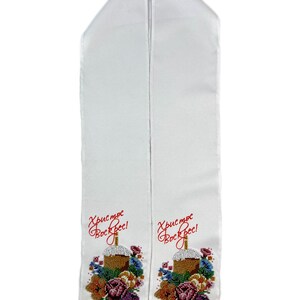 May include: White liturgical stole with embroidered floral and cake design. The stole features the text "Christ is Risen!" in red script. The embroidery includes a cake, flowers, and colorful beads.
