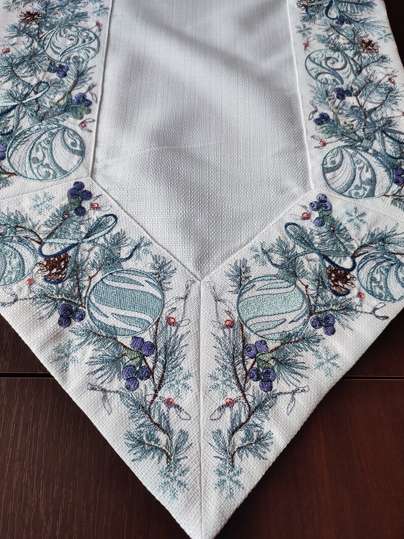以下が含まれることがあります： 青と緑の松の枝、オーナメント、ベリーが刺繍された白いテーブルランナー。デザインはランナーの4辺すべてに施されています。