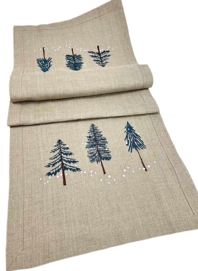 Op de afbeelding: Een beige linnen tafelloper met een geborduurd ontwerp van vijf blauwe dennenbomen en witte sneeuwvlokken.