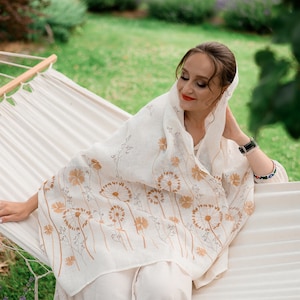 May include: A cream-colored linen shawl with a floral dandelion design in shades of brown. The shawl is draped over a person wearing a matching cream-colored outfit while relaxing on a white hammock in a garden setting.