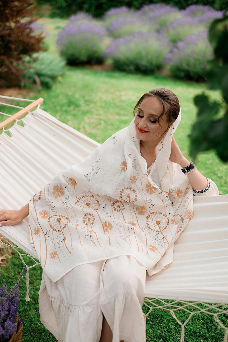 May include: A cream-colored linen shawl with a floral dandelion design in shades of brown. The shawl is draped over a person wearing a matching cream-colored outfit while relaxing on a white hammock in a garden setting.