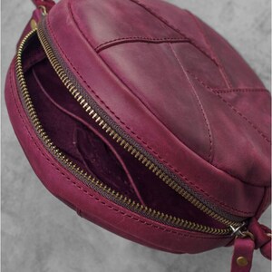 May include: A round, burgundy leather crossbody bag with a zipper closure. The bag has a stitched leaf-like design on the front and a long, thin strap. The zipper and hardware are a brass color. The bag is open, revealing the interior.