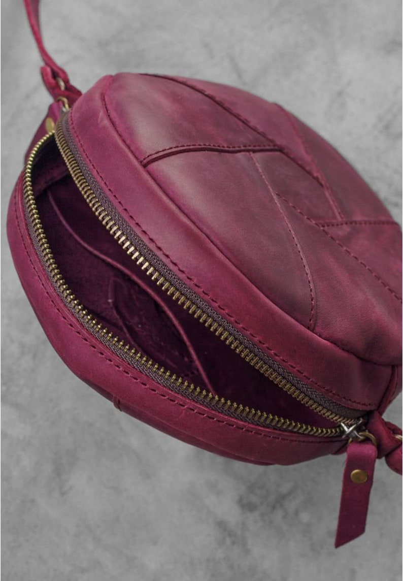 May include: A round, burgundy leather crossbody bag with a zipper closure. The bag has a stitched leaf-like design on the front and a long, thin strap. The zipper and hardware are a brass color. The bag is open, revealing the interior.