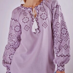 May include: A lavender blouse with a white and purple embroidered geometric pattern. The blouse has a round neckline and long sleeves with a gathered cuff.