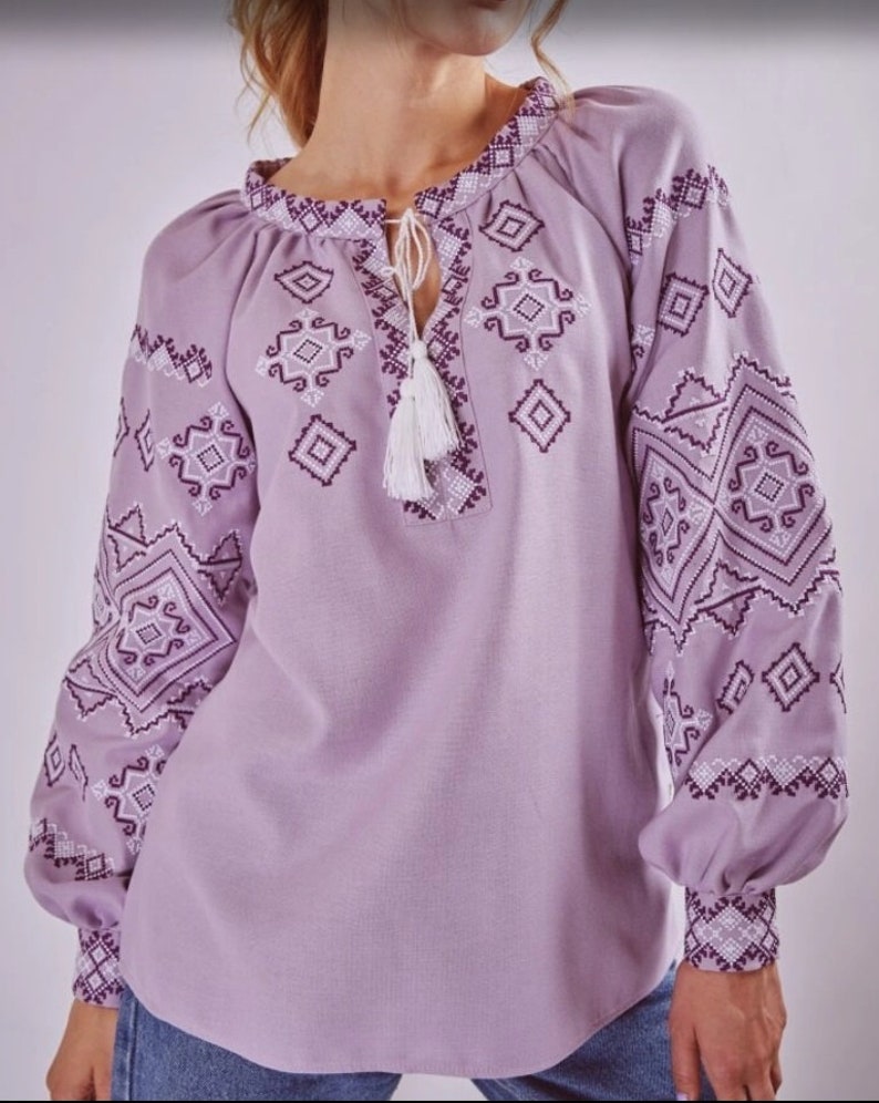 May include: A lavender blouse with a white and purple embroidered geometric pattern. The blouse has a round neckline and long sleeves with a gathered cuff.
