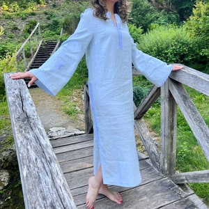 Pode incluir: Um vestido de linho azul claro com mangas compridas e largas e decote em V. O vestido apresenta detalhes bordados em um tom de azul mais claro ao longo do decote, das mangas e das costuras laterais. O vestido &eacute; comprido at&eacute; o tornozelo e tem uma fenda lateral.