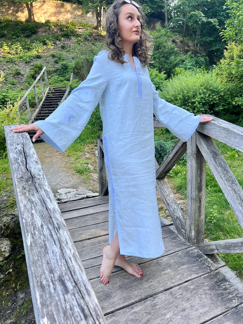 Pode incluir: Um vestido de linho azul claro com mangas compridas e largas e decote em V. O vestido apresenta detalhes bordados em um tom de azul mais claro ao longo do decote, das mangas e das costuras laterais. O vestido &eacute; comprido at&eacute; o tornozelo e tem uma fenda lateral.