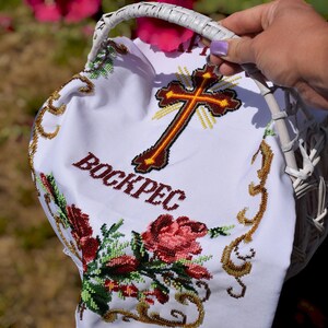 K&ouml;nnte beinhalten: Ein wei&szlig;er Weidenkorb mit einem wei&szlig;en bestickten Tuch. Das Tuch zeigt ein Kreuz, das Wort "BOCKPEC" und Blumenstickereien in Rot, Gr&uuml;n und Gold. Der Korb wird von einer Person gehalten.