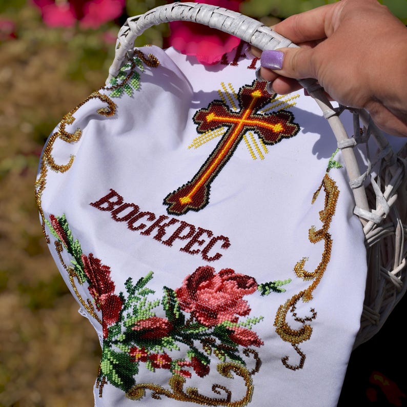 K&ouml;nnte beinhalten: Ein wei&szlig;er Weidenkorb mit einem wei&szlig;en bestickten Tuch. Das Tuch zeigt ein Kreuz, das Wort "BOCKPEC" und Blumenstickereien in Rot, Gr&uuml;n und Gold. Der Korb wird von einer Person gehalten.