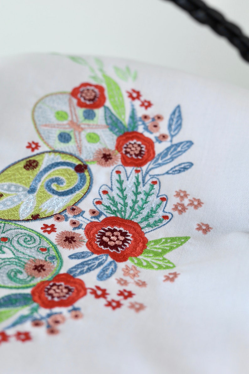 May include: Close-up of a white fabric with colorful embroidery. The embroidery features Easter egg designs in shades of green, blue, and orange, along with red and pink flowers and green leaves. The design is likely for a seasonal home decor item.