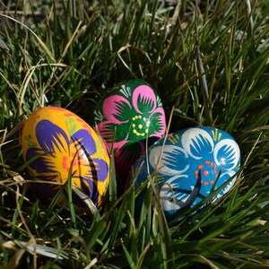 May include: Three brightly painted Easter eggs nestled in green grass. One egg is yellow with purple floral designs, another is pink and green, and the third is blue and white. The eggs are decorated with intricate floral patterns.