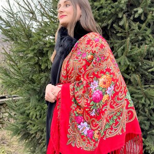 May include: A woman wearing a red shawl with a floral pattern and fringe. The shawl is draped over her shoulders and has a paisley design.