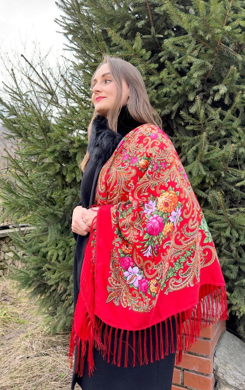 May include: A woman wearing a red shawl with a floral pattern and fringe. The shawl is draped over her shoulders and has a paisley design.