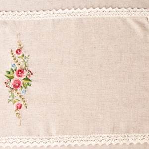 May include: Rectangular beige linen placemat with a floral embroidery design featuring pink roses, green leaves, and blue accents. The edges are trimmed with a white, scalloped lace border. A decorative textile for the dining table.