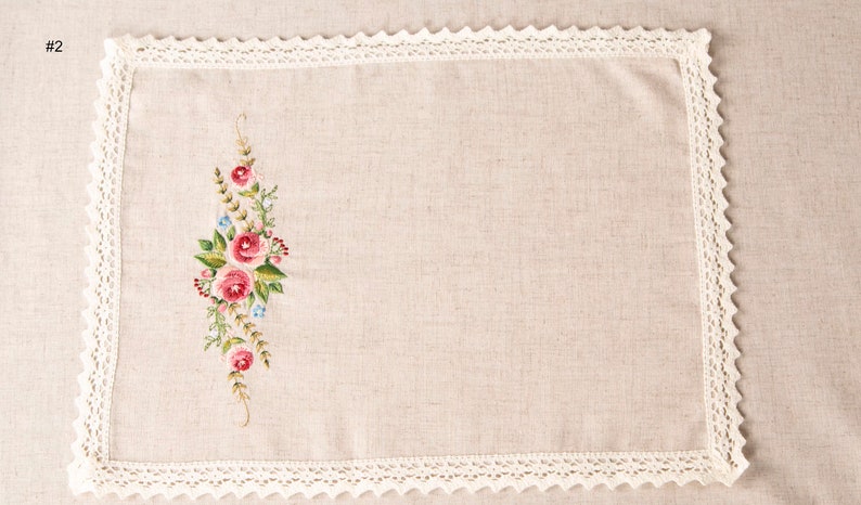May include: Rectangular beige linen placemat with a floral embroidery design featuring pink roses, green leaves, and blue accents. The edges are trimmed with a white, scalloped lace border. A decorative textile for the dining table.