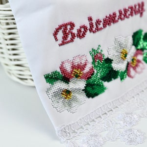K&ouml;nnte beinhalten: Wei&szlig;es besticktes Tuch mit floralen und textlichen Details. Der Text "Bożemu" ist aus roten Perlen. Das florale Design umfasst wei&szlig;e, rosa und rote Blumen mit gr&uuml;nen Bl&auml;ttern. Der untere Rand hat eine wei&szlig;e Spitzentrimmung.