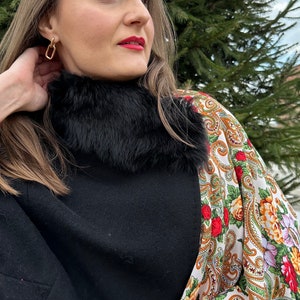 May include: A woman wearing a black coat with a black fur collar and a white and gold patterned shawl. She is wearing gold earrings.