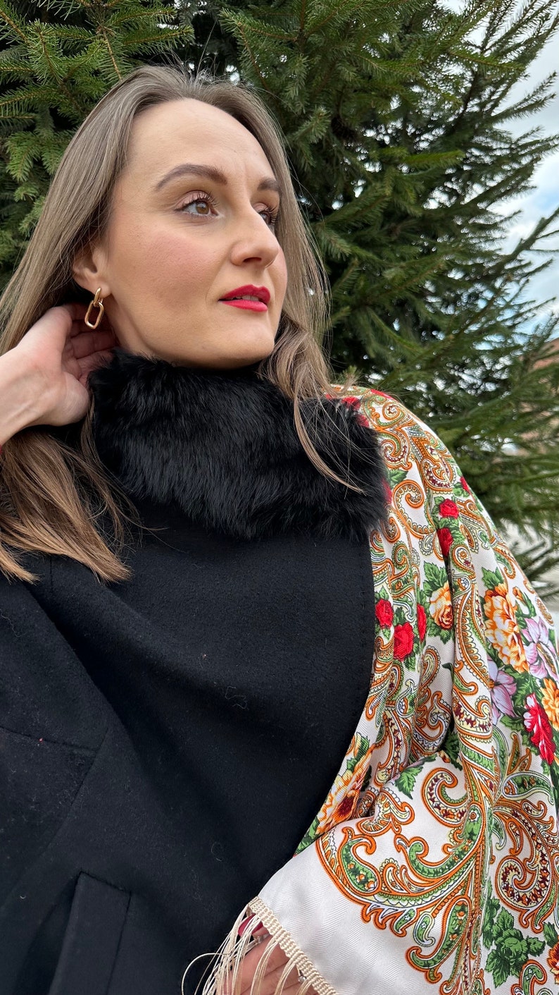 May include: A woman wearing a black coat with a black fur collar and a white and gold patterned shawl. She is wearing gold earrings.