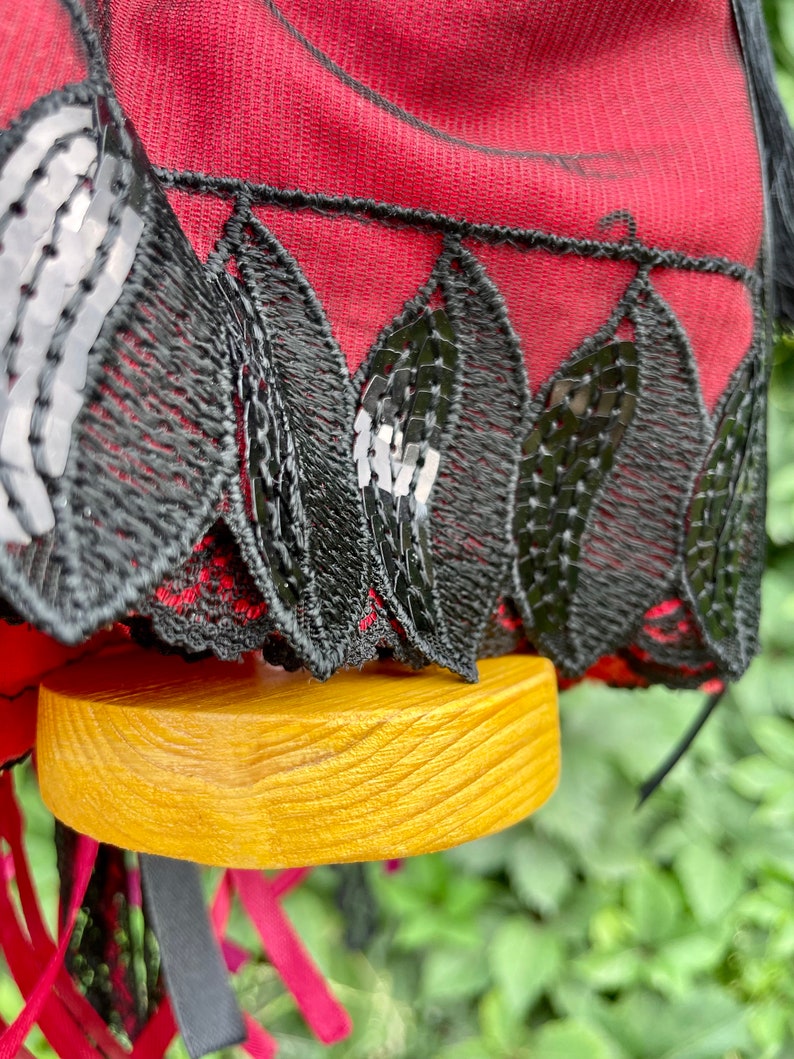 Puede incluir: Un sombrero de tela roja con ribete de encaje negro y lentejuelas. El sombrero est&aacute; sobre una base de madera.
