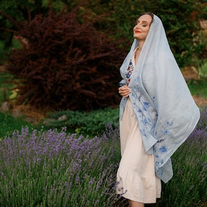 K&ouml;nnte beinhalten: Eine Frau in einem wei&szlig;en Kleid und einem hellblauen Schal mit Blumenstickerei steht auf einem Lavendelfeld.