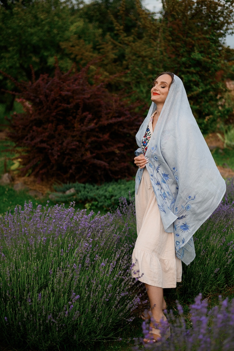K&ouml;nnte beinhalten: Eine Frau in einem wei&szlig;en Kleid und einem hellblauen Schal mit Blumenstickerei steht auf einem Lavendelfeld.