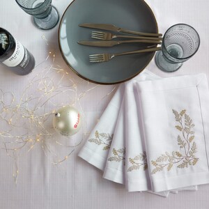 Puede incluir: Una mesa puesta con servilletas de lino blanco con dise&ntilde;os bot&aacute;nicos bordados, un plato gris con borde dorado, cubiertos y dos vasos azules texturizados. Tambi&eacute;n hay una botella de vino y una bola de Navidad.