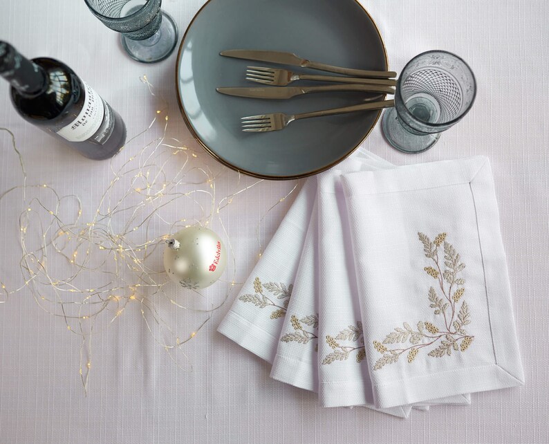 Puede incluir: Una mesa puesta con servilletas de lino blanco con dise&ntilde;os bot&aacute;nicos bordados, un plato gris con borde dorado, cubiertos y dos vasos azules texturizados. Tambi&eacute;n hay una botella de vino y una bola de Navidad.