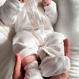 Op de afbeelding: Een witte baby romper met een kanten rand en knoopsluiting. De romper wordt gedragen door een baby met witte sokken aan de voeten.