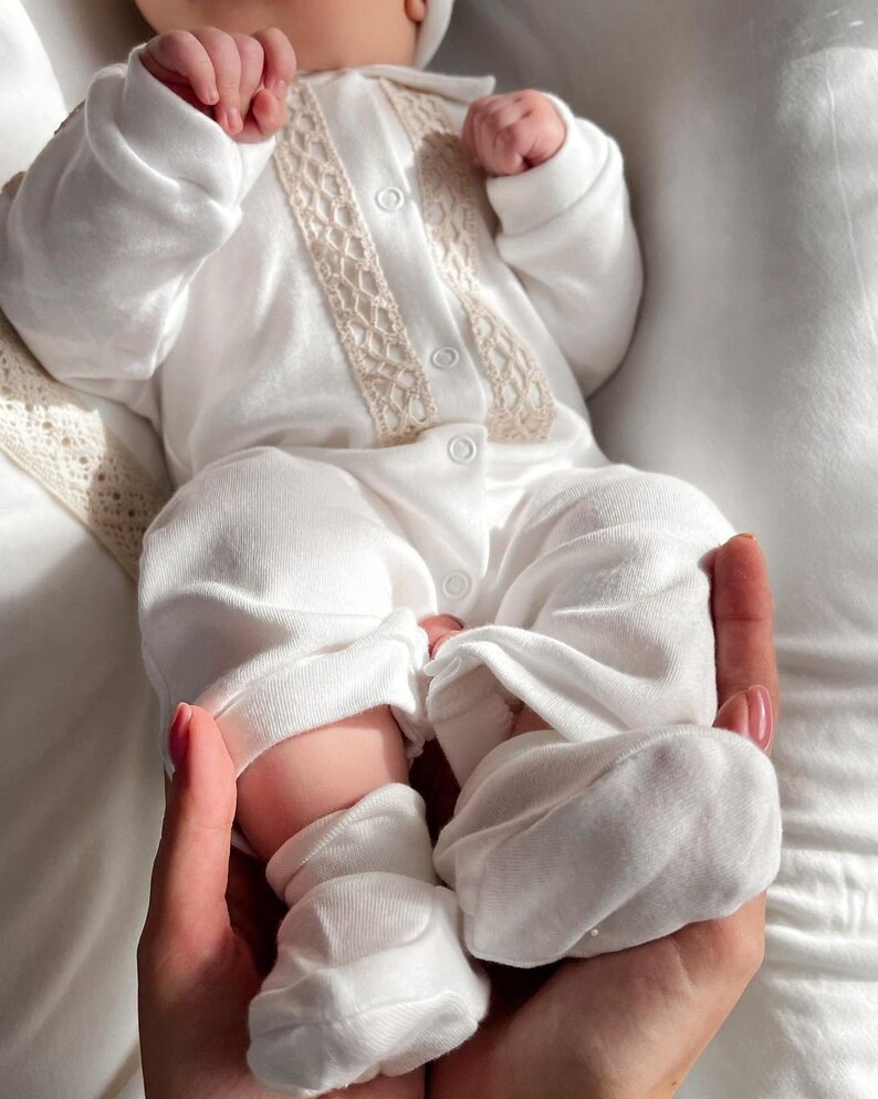 Op de afbeelding: Een witte baby romper met een kanten rand en knoopsluiting. De romper wordt gedragen door een baby met witte sokken aan de voeten.
