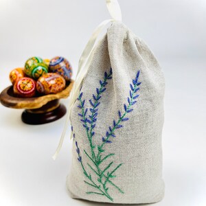 K&ouml;nnte beinhalten: Ein beiger Leinenbeutel mit Kordelverschluss, bestickt mit blauen Lavendelzweigen und gr&uuml;nen Stielen. Eine kleine Holzschale h&auml;lt im Hintergrund mehrere dekorierte Ostereier. Der Beutel ist mit einem wei&szlig;en Band gebunden.