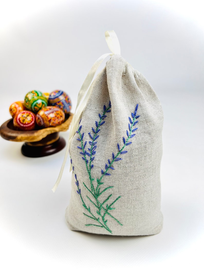 K&ouml;nnte beinhalten: Ein beiger Leinenbeutel mit Kordelverschluss, bestickt mit blauen Lavendelzweigen und gr&uuml;nen Stielen. Eine kleine Holzschale h&auml;lt im Hintergrund mehrere dekorierte Ostereier. Der Beutel ist mit einem wei&szlig;en Band gebunden.